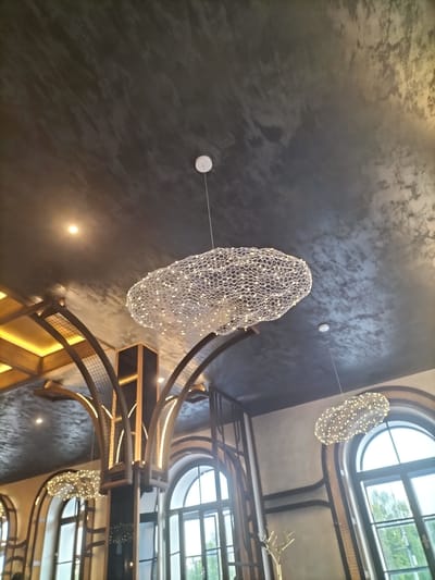 This is a low-angle, indoor shot looking up at a ceiling with unique lighting fixtures and architectural elements. The ceiling has a dark, textured finish, possibly concrete or stucco, with areas of lighter marbling. Two cloud-shaped light fixtures, composed of a mesh-like material and embedded with numerous small, warm white LED lights, are suspended from the ceiling. One larger fixture hangs prominently in the upper center of the frame, while a smaller, similar fixture is visible in the lower right.

Below the ceiling, there are decorative arched structures made of metal and wood, accented with integrated lighting that casts a warm glow. These arches suggest an interior space with a modern yet somewhat industrial or rustic design. Large arched windows are visible in the background, with panes that suggest they look out onto a green, possibly outdoor environment, hinting at daylight. The overall impression is one of a contemporary restaurant or event space. No people are visible, and there is no visible text. The time of day appears to be daytime, with natural light filtering through the windows. The overall mood is calming and aesthetically pleasing due to the design and lighting.