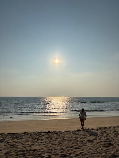 A vertical-oriented photograph depicts a coastal scene, possibly in Khok Kloi, Thailand, given the request. The left side shows a light blue sky with the sun visible. The right side features the ocean with sparkling reflections and a sandy beach. A solitary man walks on the shoreline, dressed in a light-colored shirt and shorts. The sand appears textured and dry.