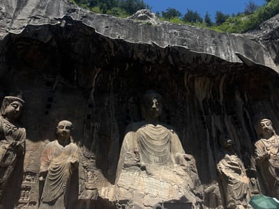 The image depicts the Longmen Grottoes, a UNESCO World Heritage Site located in Luolong District, China. The scene features several monumental Buddhist statues carved into a rocky cliff face. In the center is a large, seated Buddha figure, appearing serene and imposing. To its left and right are other figures, including what appear to be bodhisattvas and disciples, each carved with intricate details of their robes and facial features. The statues are set within natural cave-like openings, with the rough texture of the rock contrasting with the smooth finish of the carvings. Sunlight casts strong shadows across the figures, highlighting their sculptural forms and the depth of the grottoes. The top of the cliff is covered with greenery, and the sky above is clear blue.