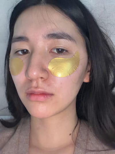 A woman is lying on a textured, light-colored surface, possibly a bed or couch. She has dark hair and is wearing gold-colored under-eye patches. A mole is visible on her neck. The scene's location is not discernible from the image.