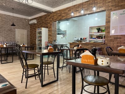 The image depicts the interior of a restaurant, likely named "Минутка" (Minutka), in Kizilyurt, Russia. The space is furnished with numerous dark-colored tables with metal frames and wooden tops, paired with matching chairs. The walls are adorned with exposed brick and tiled sections. Overhead lighting fixtures, some modern and industrial, hang from the ceiling, which has a textured finish. Various decorative elements, including a menu board on the wall and items on shelves, suggest a dining establishment. The scene is well-lit, creating a warm and inviting atmosphere. There are no visible people in the image, but the setup implies a place ready to serve customers.