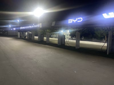 This media file captures an outdoor electric vehicle charging station at night, likely in Temirhoja, Uzbekistan. The scene is illuminated by bright, harsh overhead lights, creating strong glare and lens flare. The primary subjects are the charging stations themselves, characterized by tall, white rectangular structures housing the charging equipment. Cables are visible, suggesting the stations are ready for use.

Above the charging stations, a prominent illuminated sign displays "BYD" in large, white, futuristic lettering. To the left of the BYD logo, another illuminated sign reads "ELEKTROMOBILLAR UCHUN QUVVATLASh STANSIYASI," which translates from Uzbek to "Electric Vehicle Charging Station." This text is also in white, illuminated letters.

The ground is paved with asphalt, appearing wet in some areas, suggesting recent rain or dew. The background features trees and some fencing, indicating an urban or semi-urban environment. The overall atmosphere is functional and modern, with no people visible, suggesting the station is either closed for the night or automated. The time of day is clearly night due to the darkness and artificial lighting. The weather appears to be clear, with no indication of rain or strong wind. The lighting creates a somewhat stark and high-contrast image.