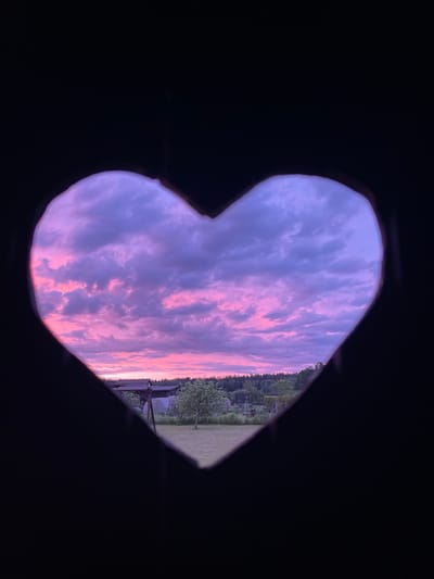 The image, taken in Zaķumuiža, Latvia, shows a heart-shaped cutout framing a twilight sky filled with dramatic clouds. The sky transitions from a deep purple at the top to vibrant pinks and oranges near the horizon, indicating either sunrise or sunset. Below the sky, a silhouetted landscape is visible, featuring the tops of trees forming a dark line against the colorful sky. A faint outline of a building or structure can be discerned within the landscape, along with hints of green grass and possibly a swing set. The foreground is dominated by the black silhouette of the heart shape, suggesting the image is viewed through a carved opening. There are no people or explicit actions depicted in the scene.