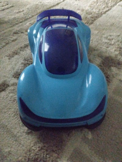 A blue toy car is placed on a beige carpet. The car has a spoiler on the back and a dark blue windshield. The car is facing towards the viewer and is the main subject of the image. The background is a textured beige carpet. The image is taken from a slightly elevated angle. There are no people visible in the image. The lighting suggests an indoor setting. There is no information available about the location in the city of Калыс-Ордо жаңы курулушу, Kyrgyzstan.