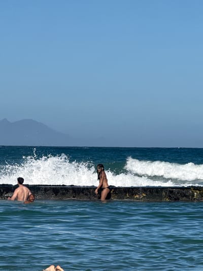 A medium shot depicts a scene at the beach, likely in Muizenberg, South Africa. A man and woman are positioned on a pier or rocky divider between crashing waves and the ocean. The man is shirtless and crouched on the pier, while the woman, wearing a dark bikini, is lying face down on the edge. A second man is waist deep in the ocean. The sky is clear and blue, with a hint of a mountain in the background.
