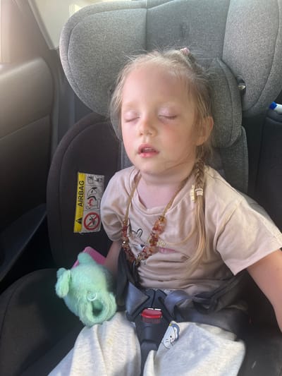 A young child with blonde hair is asleep in a gray car seat. The child is wearing a light-colored t-shirt with a graphic print, and a beaded necklace. A green plush toy is on the child's lap. The car seat has a yellow and black "AIRBAG" warning label. The background appears to be the interior of a car. This image was taken in Mal'tsevo, Russia.