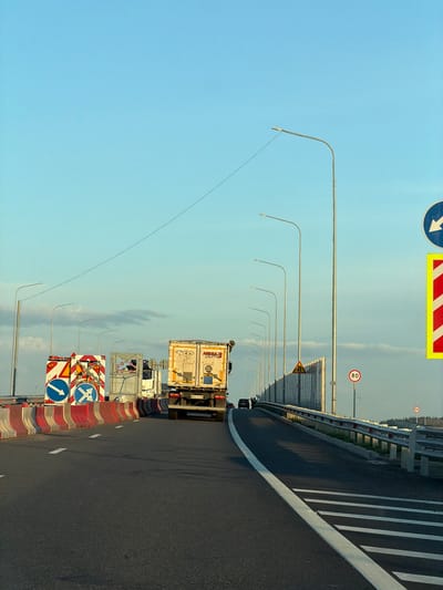 The image shows a multi-lane road with traffic, likely a highway or bridge, under a clear blue sky with scattered clouds. Several streetlights line the right side of the road. On the left, there are construction barriers made of red and white plastic. Traffic signs are visible, including directional signs and speed limit signs (80 and 60). Two large trucks are on the road, one with a white trailer and the other a white semi-truck with a white trailer. A few people are walking near the side of the road. The environment appears to be an outdoor, potentially elevated road structure. The location is identified as Zimnyaya Gorka, Russia.