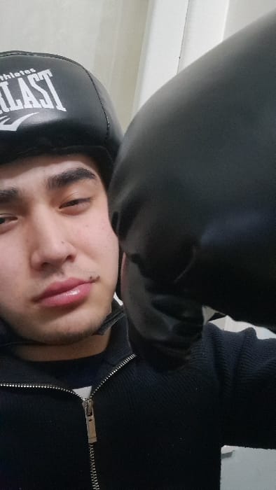 A man with a boxing headgear with the word "Everlast" is leaning towards the camera. He wears a black zip-up sweater. A black boxing glove is near his face. The background shows a white wall and a small portion of white stairs. The man has dark eyebrows, light skin, and a mole on his lip.