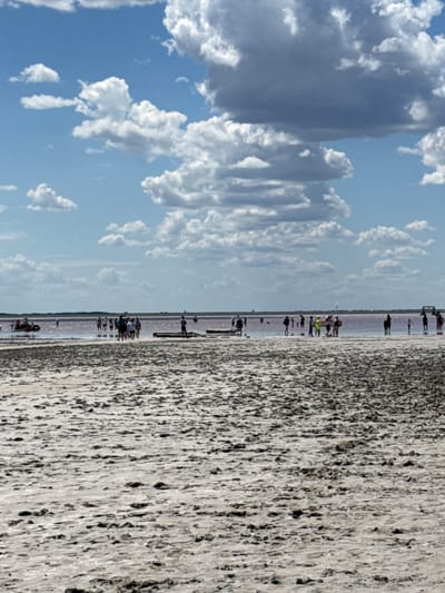 The image depicts a natural landscape with a body of water in the background and a sandy, textured foreground. Several individuals are visible in the water and along its edge, appearing to be engaged in recreational activities. Some are standing, others are walking, and a few are gathered in small groups. The water exhibits a reddish-pink hue, suggesting it might be a salt lake. The sky is partly cloudy, with a mix of white and gray clouds against a blue backdrop. The foreground consists of sand or salt flats with a rough, uneven surface, possibly indicating dried mud or salt deposits. The overall scene suggests a bright, sunny day at a natural destination. There is no specific information about the city of Бурсоль, Russia visible in the image.