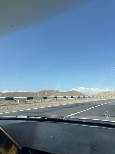 The image shows a view from inside a vehicle traveling on a paved road. The road has white lane markings and a guardrail with black and white reflective markers. To the left of the road, there are arid hills and mountains under a clear blue sky with some wispy clouds. In the distance, there appear to be contrails from airplanes. The vehicle's dashboard is visible in the foreground on the right side of the image. A Mercedes-Benz logo is visible on the road. The time of day appears to be daytime, likely late morning or afternoon, given the bright sunlight and clear sky. The weather seems to be clear and sunny. The location is Paruyr Sevak, Armenia, as indicated by the text. No people are visible in the image.