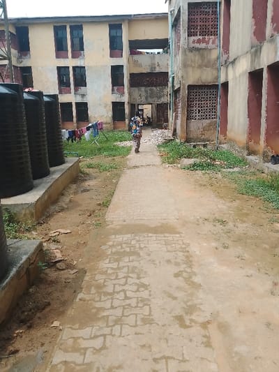 The image displays an outdoor area situated between multi-story buildings in Rumuekini, Nigeria, under an overcast sky. A light-colored paved pathway constructed from interlocking pavers traverses the scene from the foreground towards the background. To the left of the pathway, a row of five black, cylindrical water tanks rests on a concrete platform. Adjacent to the tanks and pathway, the ground consists of exposed dirt and patchy green vegetation.
In the background-left, a three-story building with a weathered light beige facade features multiple rectangular windows trimmed in dark red. A white metal clothes drying rack laden with various garments is visible on the grassy ground level in front of this building. Another line of laundry is visible on a second-story balcony.
To the right of the pathway, a two-story building with a faded beige and dark red facade presents a more rugged appearance, featuring rectangular openings and decorative concrete block screens on its upper level. Exposed piping runs vertically on its exterior.
A single individual is observed walking away from the camera, centered on the paved pathway, moving towards the background. The person is wearing a long-sleeved, brightly patterned, multi-colored shirt and light-colored trousers. Near the base of the right building, a pile of light-colored rubble is present.