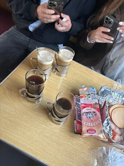 The image shows a table with four beverages in glass mugs and food items. Two of the beverages appear to be coffee with milk and the other two appear to be coffee. Food includes a foiled tray of sliced meat, a package labeled "Бобруйска" (Babruysk), and a Snickers bar. Two people are present, both holding smartphones; one is holding what appears to be money. This scene may be located in a train or other travel conveyance.
