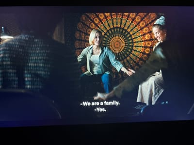 The image shows a dimly lit scene with three people. In the foreground on the left, a person is seen from the back, wearing a patterned shirt and partially obscured by shadow. In the middle, a blonde woman in a light blue shirt and jeans is seated and looking to her left with an expressive face. Behind her is a large, circular tapestry with an intricate, colorful, psychedelic pattern, predominantly featuring oranges, greens, and yellows, resembling a mandala or peacock feather design. To the right of the blonde woman, another person, possibly a young woman, is also seated and smiling, wearing a light-colored headscarf and a light-colored garment. Their hands appear to be near each other. White subtitles are visible at the bottom of the screen, displaying "-We are a family." and "-Yes." The scene appears to be indoors, with the tapestry serving as a backdrop. There is no indication of the city of Dolna Banya, Bulgaria in the image.