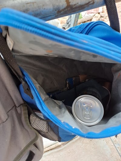 The image is a close-up, interior view of an open blue backpack with a gray outer bag attached. A silver aluminum can is visible inside the blue backpack, resting on some dark fabric. A black strap from the backpack is visible near the top left. The background is out of focus but appears to be outdoors with rocky ground and some debris. The backpack is positioned outdoors, possibly near a metal railing. The specific location in the city of Settimo Torinese, Italy, cannot be determined from the image alone.