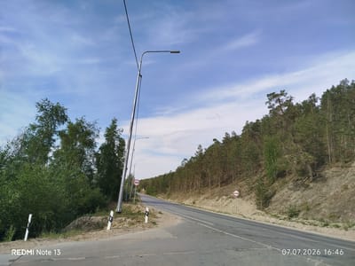 This is a medium-angle, exterior, eye-level, daytime, full-color, medium-shot photograph taken on a paved road leading through a forested area. The road is a two-lane asphalt highway with white lane markings, curving to the right as it recedes into the distance. The right side of the road is bordered by a steep, sandy embankment covered with pine trees. The left side of the road features a slightly lower, grassy area with dense green trees and vegetation.
Several objects are visible:
- Two tall, silver streetlights with horizontal lamp fixtures are positioned on the left side of the road.
- Two traffic signs are present, one on each side of the road, both displaying a circular red border with a horizontal red bar across the center, enclosing an image of two vehicles. These signs likely indicate a prohibition of certain vehicle types or overtaking.
- Two white road markers with black stripes are placed on the left side of the road, marking the edge of the embankment.
- The sky is a clear blue with thin, wispy white clouds.
A watermark in the bottom left corner reads "REDMI Note 13", and another in the bottom right corner shows the date and time "07.07.2026 16:46". The location is identified as Nizhny Bestyakh, Russia. No people or animals are present in the image. The overall scene depicts a rural or semi-rural highway environment on a clear day.