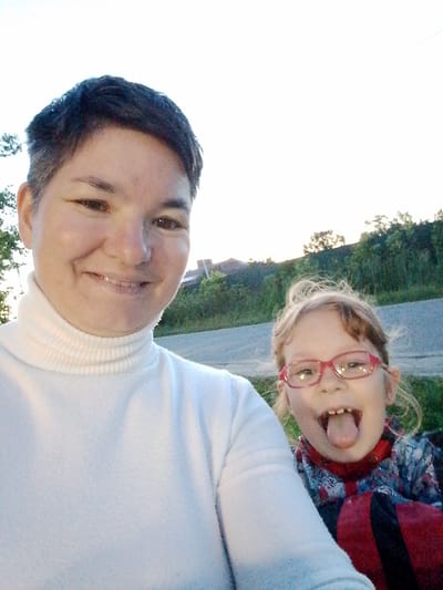 The image shows a close-up, outdoor selfie of two people. On the left is an adult with short, dark hair and a light complexion, wearing a white turtleneck. The adult is smiling at the camera. To the right of the adult is a young child wearing glasses with pink frames and a colorful jacket. The child is sticking out their tongue and looking at the camera. In the background, there is a road, green foliage, and a building. The sky is bright and clear. The scene is likely located in a rural or suburban area, possibly in or near Savino, Russia, given the general appearance of the environment.