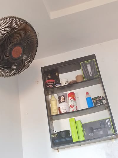 The image depicts an indoor scene, likely a bathroom or barber shop, featuring a white wall and ceiling. On the left, a dark-colored fan is mounted high on the wall. To the right, a dark metal shelf unit is affixed to the wall. The shelf unit contains various personal care and grooming products.
The top shelf holds several containers, including a red-lidded tub labeled "Vintage Hair Styling Gel," a white tub, a small brown container, and a black box. A boxed hair clipper, branded "oraimo," is also visible on this shelf.
The second shelf from the top displays a yellow bottle, a container with a picture of a person, a red and white patterned container labeled "Passion," and a blue bottle. A box labeled "Dexe A wash Black" is also present.
The bottom shelf contains a black bowl or basin, two green boxes, and a large black box containing what appears to be a hairdryer, also branded "oraimo." The overall impression is of a functional space stocked with hair care items. The specific location in Abeere, Nigeria, cannot be determined from the visual information provided.