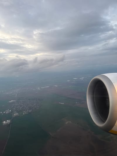 An eye-level, outdoor shot shows the sky with many clouds on the left, the land on the right, and part of a plane engine above. The city of Vecsés, Hungary can be seen in the center of the image. The plane is likely above the city, capturing a unique aerial perspective of the area. The shot appears to be taken from a passenger seat of the plane, with the engine in full view.