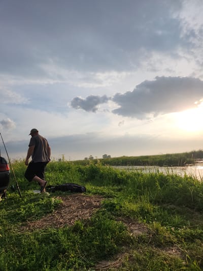 This media file captures a person fishing by a body of water, likely a canal or river, in Atameken, Kazakhstan. The scene is set outdoors during what appears to be late afternoon or early evening, as indicated by the soft, diffused sunlight breaking through the clouds. The sky is filled with a mix of light and dark clouds, suggesting a dynamic weather pattern.

The main subject is a person, seen from the back, wearing a grey t-shirt, black shorts, and a baseball cap. They are crouched down near the edge of the water, facing away from the camera. To their right, a black bag rests on the grassy bank, and several fishing rods are visible, planted in the ground or propped up. The ground is covered in lush green grass, with some dry, straw-like material also present. The water's edge is lined with dense green vegetation, which continues to the horizon. The overall setting is natural and serene, with a focus on the solitary activity of fishing. There are no visible text elements in the image.