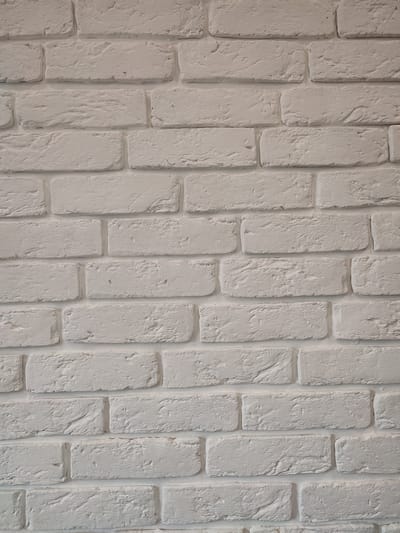 The image shows a close-up, full-frame view of a white brick wall. The bricks are rectangular and laid in a staggered pattern, typical of traditional brickwork. The surface of the bricks has a textured, somewhat rough appearance, with visible mortar lines between them. The lighting appears to be even, suggesting an indoor setting or overcast conditions, with no strong shadows. The color is a uniform off-white or very light grey. There are no people, animals, or other objects visible in the frame. The perspective is straight-on, with the camera positioned perpendicular to the wall. The composition is simple and focused solely on the texture and pattern of the brickwork. The scene does not provide any information that would allow for the identification of a specific location within Ivanovska, Russia, or any other geographical area.