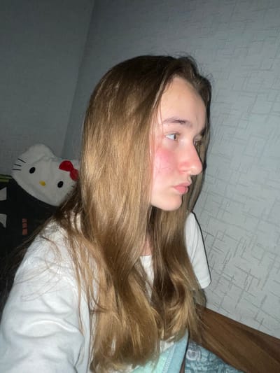 A medium shot photograph captures a young person with long, light brown hair, seen in profile from the right side of their face. They are wearing a white t-shirt and have noticeable red flushing on their cheeks. The background consists of a textured gray wall and a wooden surface along the bottom right of the frame. To the left, a white plush toy resembling Hello Kitty with a red bow is visible. The scene appears to be indoors, likely in a residential setting in Novopillia, Ukraine.