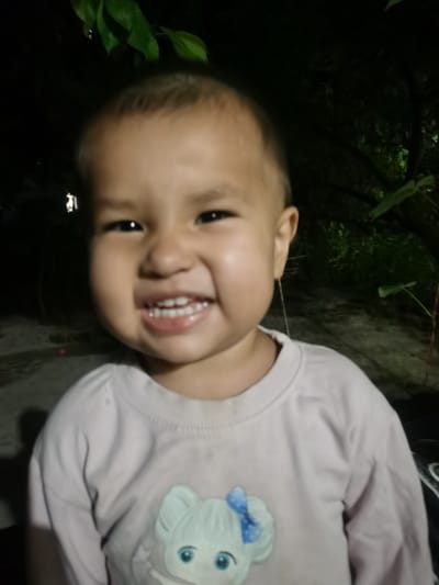 A child, likely under the age of five, with fair skin and dark eyes, is smiling broadly, revealing their teeth. They are wearing a light-colored long-sleeved shirt with a cartoon image of a character with blue eyes and a blue bow on the chest. The background is dark, with out-of-focus foliage suggesting an outdoor setting at night, possibly in Karabair, Uzbekistan. The lighting appears to be artificial, casting shadows and highlighting the child's face.