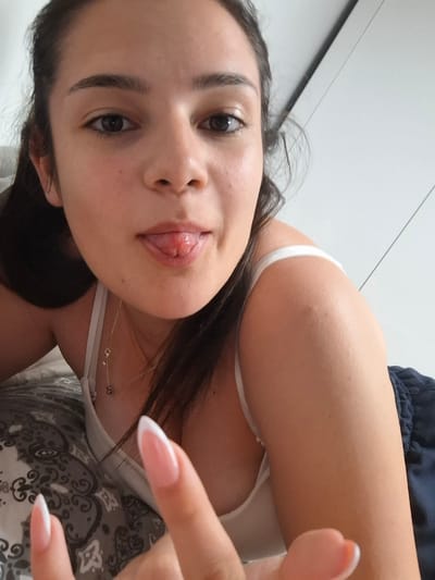 This image captures a close-up, eye-level indoor scene in the city of Choba, Bulgaria, featuring a young adult female looking directly at the camera. The subject has dark hair pulled back, visible dark eyes, and a light complexion. She is sticking out her tongue, which exhibits a reddish, raised papilla or lesion near the tip. Her left hand is partially visible in the lower foreground, raised towards the camera, displaying long, almond-shaped nails with a white French tip design. She wears a white spaghetti strap top, revealing part of her chest and shoulder, and what appears to be dark blue, possibly satin, shorts or pajama bottoms. A delicate silver-toned necklace with small charms is visible around her neck. She is lying on a surface covered with a grey and white patterned textile, likely a bedspread or pillowcase, featuring ornate designs. The background consists of a plain white wall or furniture, possibly a wardrobe, with visible vertical and diagonal seams or panel lines. The lighting is soft and even.