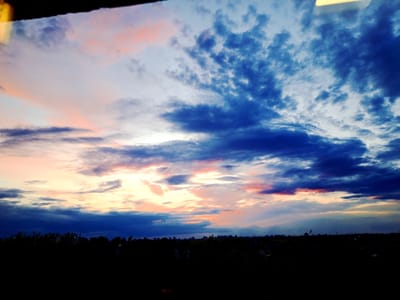 A dramatic, wide-angle shot captures a vibrant sunset over a silhouetted landscape, likely in the vicinity of Сеніцкі сельскі Савет, Belarus. The sky is a canvas of intense color, with deep blues dominating the upper portion, transitioning to lighter blues and whites closer to the horizon. Wispy clouds are painted with strokes of soft pink and orange, reflecting the last rays of the setting sun. The lower third of the image is consumed by a dark, dense silhouette of trees and what appears to be low-rise buildings, creating a stark contrast against the illuminated sky. The overall impression is one of a serene, yet powerful natural spectacle.