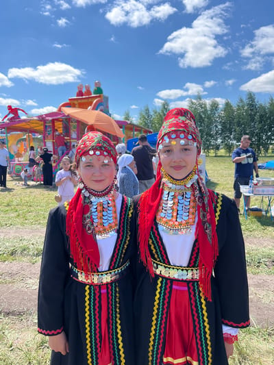 The image depicts a sunny outdoor scene in Staraya Kosa, Russia, likely at a festival or fair. Two young girls, dressed in traditional, brightly colored folk costumes, are the primary subjects, standing close together and looking at the camera. Their attire features red headscarves with intricate patterns, layered necklaces adorned with numerous silver coins and colorful beads, and black tunics over red underskirts with decorative trim. In the background, a lively atmosphere is evident with a large, colorful inflatable playground structure, a crowd of people of various ages milling about, and a table set up with food or supplies. The overall setting suggests a community gathering on a clear, blue-sky day.