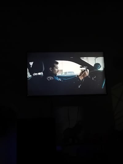A screen, situated within a dimly lit room in Timaná, Colombia, displays a still image depicting a male police officer in the driver's seat of a vehicle. The officer, with a mustache and dark hair, wears a dark blue uniform shirt featuring a circular emblem on his left shoulder. His hands are placed on the steering wheel. Inside the vehicle, a dark headrest is visible behind him. Mounted on the dashboard or windshield area are a small, dark cylindrical device and a larger, dark, round object, consistent with vehicle-mounted equipment. Through the vehicle's windshield, a daytime urban or suburban environment is visible, characterized by light-colored structures and the partial presence of another vehicle in the background. The immediate surroundings of the screen are dark, with a faint blue light emanating from the lower left foreground.