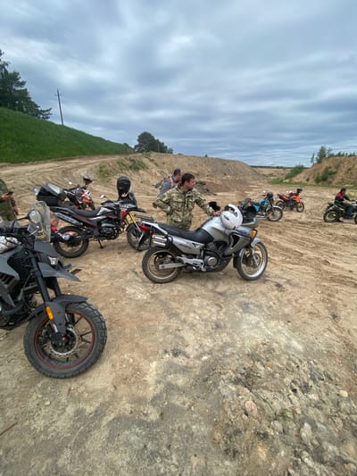 The image depicts a group of motorcycles parked on a dirt road or terrain, likely in a rural or undeveloped area of Верцялішкаўскі сельскі Савет, Belarus. Several individuals are present, some wearing military-style camouflage clothing. They appear to be examining or working on one of the motorcycles. The environment consists of a dirt or sandy ground surface, a grassy slope on one side, and a higher dirt embankment in the background. The sky is overcast with cloudy conditions.