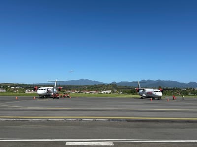 Two turboprop aircraft are parked on an airport tarmac under a clear blue sky. The aircraft on the left is white with red and black accents, and the word "Wings" is visible in red lettering on its fuselage. It is flanked by two orange traffic cones and appears to be undergoing service, with a small utility vehicle and several individuals in orange vests standing nearby. The aircraft on the right is also white with red and black accents, and the word "Wings" is visible in red lettering on its fuselage. It is situated further down the tarmac, and two individuals in orange vests are visible near it. The tarmac is marked with yellow and white lines. In the background, there is a strip of green grass, followed by a line of houses with red roofs and palm trees, and beyond that, a range of rolling, tree-covered hills and mountains. The scene is a typical view of an airport in a tropical or subtropical location. The location is the Lempeh Airport (LMP) in Lempeh, Indonesia, based on the distinctive landscape and vegetation.