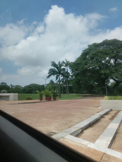 This is a photograph taken from a vehicle, looking out onto an open paved area in front of a park. The paved area is a light reddish-brown color and is bordered by concrete and stone structures. To the left, a portion of a white concrete building or wall is visible. In the mid-ground, there are three potted plants lined up on the pavement, with palm trees and other lush greenery behind them. The background features a dense forest of trees, including a very large, mature tree on the right side of the frame. The sky above is partly cloudy with a blue sky peeking through. The overall impression is one of a well-maintained park or garden area in a tropical or sub-tropical environment. The image location is Onibudo, Nigeria.