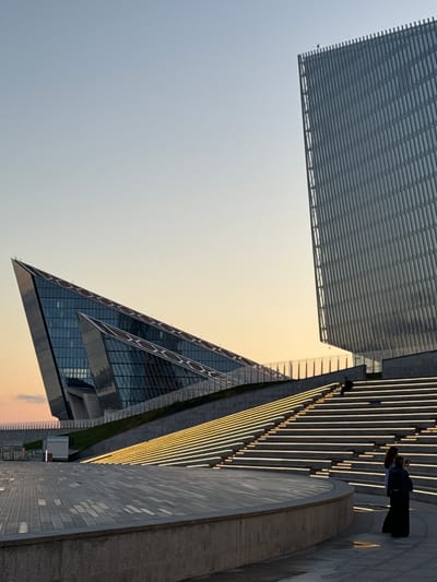 The image captures a modern architectural complex in the city of Alyumino, Russia. Two prominent buildings dominate the scene: a geometrically striking, angular structure with a glass facade, and a taller, rectangular building with a textured, vertical facade. The sky is a clear, pale blue, transitioning to a soft orange hue near the horizon, suggesting either sunrise or sunset.

In the foreground, a wide, paved plaza leads to a grand, illuminated staircase that ascends towards the buildings. The steps are lined with bright, horizontal lights, creating a warm glow. Two individuals are visible on the lower part of the plaza. One person, dressed in dark clothing, appears to be taking a photograph with a phone. Another person stands beside them, facing away from the camera. Further up the illuminated staircase, another lone figure is seated, silhouetted against the lights. The overall impression is one of contemporary design and serene outdoor space.