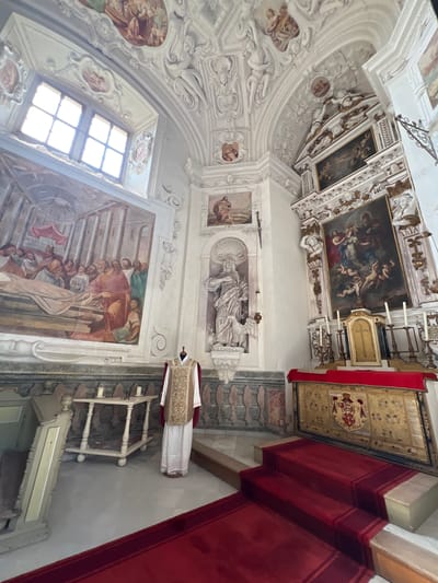 The image depicts the interior of a richly decorated church, likely a chapel, featuring ornate stucco work on the vaulted ceiling and walls, adorned with frescoes and paintings. In the foreground, a mannequin is dressed in a white alb and a gold and white chasuble, a liturgical vestment. To the left, a large fresco covers a significant portion of the wall, depicting a scene with numerous figures, a bier, and architectural elements. A wooden table with two candles stands in front of the fresco. To the right, an altar is draped with a red cloth, and behind it, a large ornate tapestry with a coat of arms is displayed. Steps covered in red carpet lead up to the altar. A statue of a saint, possibly John the Baptist given the inscription "IOANNES" visible below it, stands in a niche to the left of the altar. The overall impression is one of solemnity and historical grandeur. This scene is located in the Náchod Castle in Náchod, Czechia, specifically within the Castle Church of Saint Peter and Saint Paul.