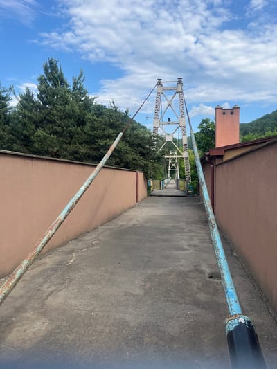 An outdoor scene captured in Kamianytsia, Ukraine, features a pedestrian suspension bridge stretching across the center of the frame, viewed from an elevated paved pathway. The bridge is constructed with distinctive A-frame metal towers and a truss-like structure painted white or light grey, showing signs of weathering. On the vertical members of the bridge's framework, the word "ZAKARPATTYA" is visibly repeated in white text. The bridge deck appears to be made of wood or a similar material, with visible handrails.
To the right of the paved pathway, a building with a light brown/tan wall runs parallel to the path. A rusty, light blue metal pipe or railing extends along the edge of this path, separating it from the building. In the distance, behind the bridge and to the left, another building with a reddish-brown section and a chimney-like structure is visible.
The surrounding environment is lush with greenery, including dense conifer trees and bushes, particularly on the left side of the frame and around the base of the bridge. Rolling green hills covered in dense foliage can be seen in the far background. The sky is bright blue with scattered white clouds, indicating clear weather and daytime. No people or specific activities are depicted in the image, suggesting a tranquil and static scene.