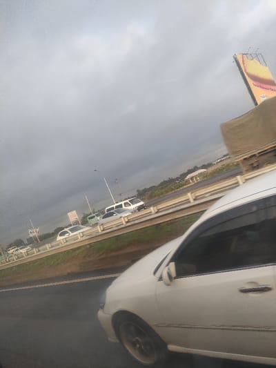 The image captures an overcast daytime scene on a multi-lane highway within Murera ward, Kenya, viewed from the interior of a moving vehicle. In the immediate foreground, the right side profile of a white sedan with gold-colored multi-spoke wheels is visible, occupying a paved road lane. The vehicle appears to be in motion.

Across a central median barrier, composed of white concrete and metal guardrails, traffic is observed traveling in the opposing direction. This includes a white passenger van, a light-colored sedan, and other vehicles in the distance. Multiple streetlights are erected along the median.

The background features dense green vegetation lining the far side of the highway, with scattered residential or commercial structures visible through gaps in the foliage. A large, angled billboard on the far right displays a partially legible advertisement, with text including "Travle Manyuazi Beds." A smaller, square billboard is also present further down the highway on the left side of the opposing lanes. The sky is uniformly gray and cloudy.