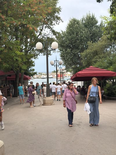 This is a candid, eye-level, full-shot photograph taken on a bright day in the city of Krk, Croatia. In the foreground, two women are walking towards the camera on a paved walkway lined with trees on the left and a covered seating area with red awnings on the right. The woman on the left is wearing a pink and white striped shirt and dark pants, and she is holding her hand to her face. The woman on the right is wearing a blue tank top and wide-legged blue pants and is carrying a white cloth. In the background, several other people are walking and standing around. Beyond the walkway, a body of water with several boats is visible, and further in the distance, there are buildings. The overall atmosphere is casual and leisurely, suggesting a tourist area.