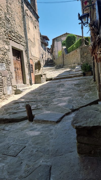 This is a low-angle, vertical, eye-level shot of a steep, cobblestone street in the historic town of Rupit, Spain. The stone buildings lining the street are old, with rough-hewn stone walls and weathered plaster. Wooden doors with iron details are visible on the left. Sunlight casts sharp shadows across the uneven stone pavement. The street curves and ascends, disappearing into the distance. A stone wall with a manicured hedge is on the right, leading to a traditional streetlamp. Dried corn husks hang to the right of the lamp. The sky is clear blue. The scene depicts a quiet, ancient village street, with no people present.