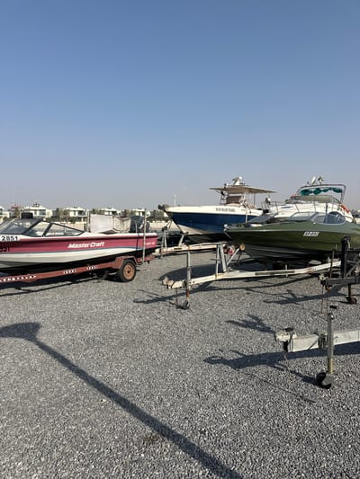 The image depicts an outdoor scene at what appears to be a boat storage area, possibly near Al Jazeerah Heritage Village in the United Arab Emirates, given the architectural style of the buildings in the background. The ground is covered in a layer of small, gray gravel. In the foreground and mid-ground, several boats are parked on trailers.
The leftmost boat is a red and white speed boat with "MasterCraft" branding and the number "2851" visible. It is on a red trailer with visible wheels and towing mechanism. To the right of this boat, there is a blue and white boat on a trailer with "ECO BLUE TOURS" written on its side. Further to the right, a dark green boat with the license plate "AP-8192" is positioned on a trailer. Other boat trailers, some empty and some possibly for smaller vessels, are also visible.
In the background, modern white buildings with flat roofs and palm trees are discernible, suggesting a developed coastal or near-coastal area. The sky is clear and bright blue, indicating a sunny day. There are no people visible in the image, and no actions or interactions are taking place. The overall impression is one of a storage facility for recreational watercraft.