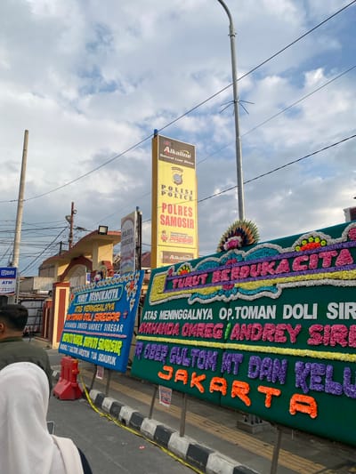 The image depicts an outdoor scene in Pasar Pangururan, Indonesia. In the foreground, two individuals are partially visible from behind, one wearing a white hijab and the other a dark jacket. They are standing on a sidewalk next to a street. In front of them are several large floral arrangements, commonly known as "papan bunga" or funeral wreaths, which are a cultural tradition in Indonesia. These wreaths are elaborately decorated with colorful flowers and text.

One prominent wreath, a deep blue, displays the text "TURUT BERDUKACITA ATAS MENINGGALNYA OP.TOMAN DOLIDIR SGAR AYAHANDA OMREG / ANDREY SIREGAR" and further down "SUPARTU/MAJOL, BUPATI SAMOSIR VAN DOKOT GULTOM / ARSTON TUA SIDARUR". The text is in white and yellow, with some words outlined in pink. The wreath is adorned with a peacock feather decoration.

Another large green wreath to the right also carries text that is partially obscured but reads "ATAS MENINGGALNYA OP. TOMAN DOLI SIR" and beneath it "AYAHANDA OMREG / ANDREY SIREGA". Below this, the text appears to be "P OBER GULTOM MT DAN KELU". The last word visible is "JAKARTA". The letters on this wreath are in vibrant purple and orange.

Behind the wreaths, a yellow sign is visible with "Alkaline Baterai Luar Biasa" printed on it. Further in the background, there is a building with an arched entrance and signs that read "POLRES SAMOSIR". A street lamp is also present, along with power lines and a cloudy sky. The presence of yellow caution tape on the ground suggests a controlled or restricted area. The overall scene suggests a public gathering, possibly a funeral procession or memorial service.