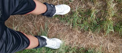 An overhead perspective captures the lower extremities of an individual standing on an outdoor surface in Weddagala, Sri Lanka. The subject wears black athletic shorts with a thin red trim visible at the leg openings. Black fabric ankle weights are secured around both ankles, positioned above white athletic shoes with white laces. Visible text on the left ankle weight indicates "1kg". The ground consists of uneven brown soil interspersed with dry, light brown grass and scattered patches of green grass. The vegetation is sparse in some areas, revealing more exposed earth.