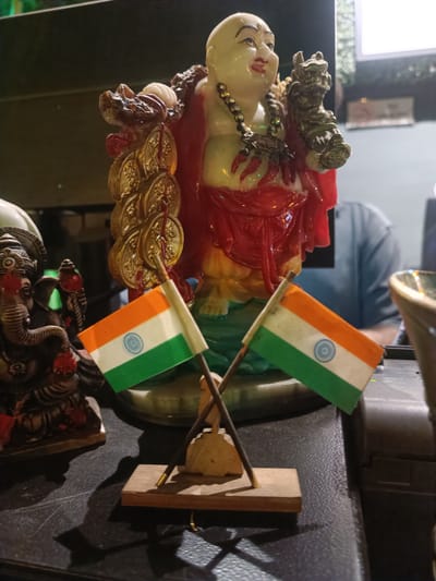 The media file showcases a collection of decorative objects placed on a dark, reflective surface, likely a table or shelf. In the foreground, two small Indian flags are prominently displayed, crossed and mounted on a simple wooden stand. The flags, with their characteristic tricolor of saffron, white, and green, and the Ashoka Chakra in the center, are a clear representation of India.
Behind the flags, a statue of a jovial, rotund figure, likely a Laughing Buddha, is a central element. This figure is adorned with a red robe, a beaded necklace, and appears to be holding a string of gold coins in its left arm and a dragon-like ornament in its right. To the left of the Laughing Buddha, a dark statue of Lord Ganesha, the elephant-headed Hindu deity, is partially visible.
The background is somewhat blurred, suggesting a shallow depth of field. It reveals hints of a person in a light blue shirt, seated behind the statues. The setting appears to be indoors, possibly a shop or a home, with some lighting fixtures visible in the upper right portion of the frame. The overall impression is one of cultural and religious symbolism, with the Indian flags suggesting a national or patriotic theme. The lighting suggests it might be evening or an indoor setting with artificial light. The presence of the Laughing Buddha and Lord Ganesha statues indicates a context where such religious and cultural iconography is displayed. No visible text is present in the image.