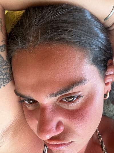The image shows a close-up of a woman's face. The woman is lying on her back and looking down at the camera with a neutral expression. Her hair is parted to the side and brushed away from her face. The woman has dark brown hair, dark brown eyes, and a light-colored complexion. Her left eye is slightly closed. Her skin is flushed, possibly due to sun exposure. She has a small, silver stud piercing in her nose. The woman is wearing a black and white patterned shirt. Her left arm is visible, and it has a tattoo of a black design. The image is taken at an angle, likely from below the woman's face. This photo was taken at the coordinates -1.825, -80.754, which is located in Montañita, Ecuador. Montañita is a popular tourist destination known for its beaches and nightlife.