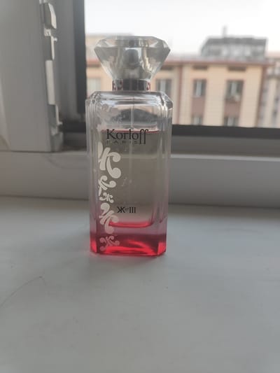 A clear glass perfume bottle with a faceted crystal cap sits on a white windowsill. The bottle contains a pink liquid and has decorative white swirls and the text "Korloff PARIS" printed on it. In the background, blurred buildings are visible through a window. The scene is located in the city of Kyngyr, Uzbekistan.