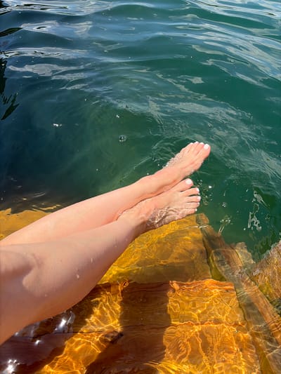 In Novomykolaivka, Ukraine, on a wooden surface partially submerged in water, two legs and feet with white nail polish are visible. The legs extend from the left side of the frame towards the center, with one leg positioned in front of the other. The feet are immersed in clear water, creating ripples and distortions on the wooden surface below. The water itself is a deep green hue, with sunlight reflecting off its surface and the submerged wood, creating patterns of light and shadow. The scene appears to be a serene moment of relaxation by a body of water.