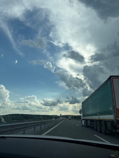 An overcast sky with scattered clouds is visible above a highway with a large semi-truck in the right lane. The highway is lined with a metal guardrail and has white lane markings. In the distance, rolling hills covered in vegetation can be seen. The image appears to have been taken from the perspective of a passenger in a vehicle traveling on the highway. There are no people or buildings visible in the frame. The location is described as Yazdach, Bulgaria.