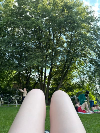 The image captures a sunny outdoor scene in Gronau, Germany, likely during the summer. The foreground is dominated by a close-up view of a person's legs, suggesting the photo is taken from a reclining position. In the background, a lush green lawn serves as the setting. Several people are visible enjoying the day. To the left, a man is using an outdoor exercise machine. Further back, near a set of outdoor gym equipment with a green towel draped over it, two other individuals are relaxing on towels on the grass. The scene is framed by a large, leafy tree canopy overhead, with patches of blue sky and white clouds visible through the foliage. The overall atmosphere is one of leisure and recreation in a park-like environment.