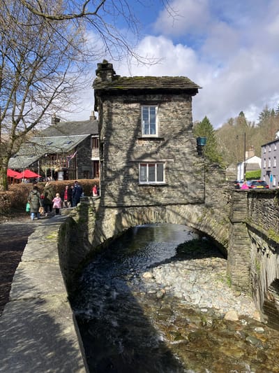 The image displays the historic Bridge House in Ambleside, Cumbria, United Kingdom, situated directly over the Stock Ghyll stream. The two-story structure is constructed of local grey stone and slate, featuring a moss-covered slate roof with a stone chimney. Two white-framed, multi-pane windows are visible on the building's facade, one on each floor.

The Stock Ghyll flows from the left foreground towards the right, passing under a single stone arch supporting the Bridge House. The stream bed contains numerous river stones and pebbles, with clear, flowing water. A stone retaining wall borders the stream's left bank, above which is a dirt path partially covered by shadows from bare tree branches. On this path, a group of approximately eight to ten individuals, dressed in winter coats and jackets, are seen walking or standing. Behind them, a larger, multi-story stone building with a slate roof, prominent windows, and outdoor seating areas with red umbrellas and a Union Jack-patterned bunting is visible.

To the right of the Bridge House, a paved road runs parallel to the stream, separated by a low stone wall. A dark grey Audi and at least one other vehicle are visible parked or stopped on this road. Further along the street, traditional stone buildings with slate roofs line the right side of the road.

The background consists of a treed hillside featuring both bare deciduous and green coniferous trees. The sky is bright blue with scattered white cumulus clouds, indicating a sunny day. Sunlight casts distinct shadows from the trees and the Bridge House across the path and the stream.