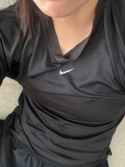 The image displays a close-up, top-down perspective of an individual's upper torso and neck area, located in Santa Catarina, Mexico. The subject is wearing a black, long-sleeved athletic shirt with a V-neck. A silver Nike "swoosh" logo is visible on the left chest of the shirt. The text "DRI-FIT" is vertically printed in white on the lower left side of the garment. A thin, silver-colored chain necklace is present around the subject's neck. The upper portion of the frame shows the subject's chin and lower lip, with dark brown hair visible on the left side of the neck/shoulder area. The background consists of a light-colored, textured surface. No specific actions or interactions are depicted.